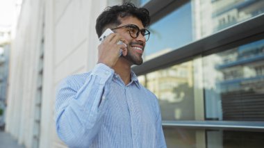 Sakallı genç adam şehir merkezinde akıllı telefondan konuşuyor. Güneşli bir günde şehir caddesinde yürürken gülümsüyor ve gözlük takıyor..