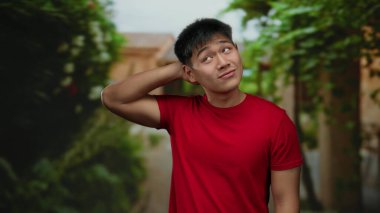 Kırmızı tişörtlü genç bir adam parkta dikiliyor. Etrafı yemyeşil yemyeşil, açık havada huzurlu bir günü simgeliyor..