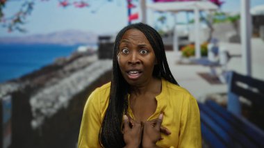 Sarı tişörtlü, uzun saçlı bir kadın güneşli bir günde deniz manzaralı bir sahilde dikiliyor..