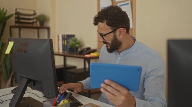 Ofiste tablet ve bilgisayar kullanan sakallı İspanyol bir adam, odaklanmış çalışma ortamı için bitkiler, kitaplar ve kırtasiye malzemeleriyle çevrili..