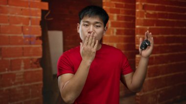 İnşaat alanında öpüşen genç bir adam, elinde anahtarlar, kırmızı gömlek, tuğla duvar zeminine yaslanmış, sanayi ortamında sevgisini ifade ediyor..