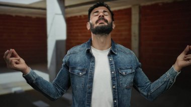 İspanyol bir adam inşaat alanında gözleri kapalı meditasyon yapıyor kot ceket giyiyor, ham endüstriyel ortamda rahatlık ve farkındalık taşıyor..