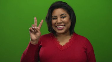 Kırmızı süveter giyen, yeşil arka planda barış işareti gösteren bir kadın pozitif ve canlı bir ortamda neşe ve güven ifade ediyor..