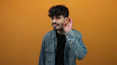 Koyu renk saçlı ve kot ceketli genç adam turuncu bir arkaplanda tuhaf bir ifade sergiliyor..