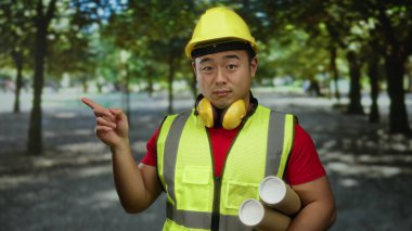 Parkta inşaat işçisi elinde bina düzenini gösteren güvenlik kaskı takan planlar tutuyor.