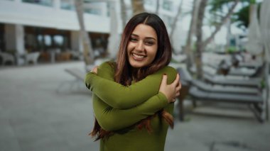 Güneşli bir günde ağaçlar ve arka planda sıcaklığı ve mutluluğu tasvir eden insanlarla dolu, açık havada gülümseyen bir kadın.