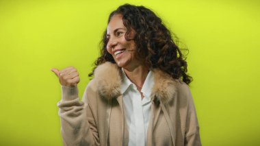 Palto giymiş kıvırcık saçlı, canlı sarı arka planda gülümseyen ve yanlamasına bakan İspanyol kadın..