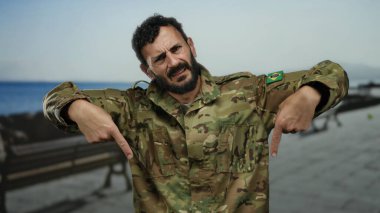 İspanyol üniformalı bir adam kumsalda geziniyor, ciddi bir ifadeyle aşağıya bakıyor, arkasında bir kıyı şeridi olduğunu düşündüren okyanus var..