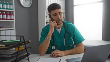 Hastane odasında çalışan İspanyol bir adam steteskop ve dizüstü bilgisayarlı telefon kullanarak klinik çalışma alanını iş günü boyunca kapalı tutarak profesyonel odaklanma ve niyetle gösteriyor.