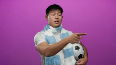 İzole edilmiş pembe bir arka planda futbol topunu tutan ve açık mavi eşarp takan genç adam ciddi bir ifadeyle işaret ediyor..