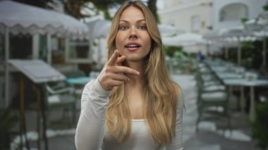 Kafkasyalı sarışın kadın restoranın terasında kamerayı işaret ediyor; sürpriz davetiye ile heyecan nişanı.