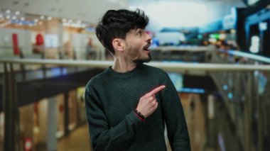 Genç adam, hareketli bir alışveriş merkezini işaret ederek canlı bir alışveriş havası ve şaşkınlık ve merak ifadeleri sergiliyor..
