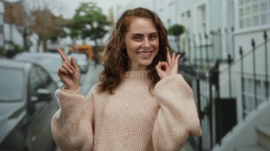 Gülümseyen ve işaret eden genç bir kadın şehir sokaklarında pembe bir süveter giyerek mutlu bir şehir ortamında dikilirken arka planda arabalarla sokak manzarası seyrediyor..