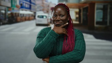 Şehir caddesinde kendine güvenen bir kadın yeşil süveter ve pembe örgüler takıyor. Etrafı şehir hayatı ve araçlarla çevrili. Açık havada güven ve şehir enerjisi taşıyor..