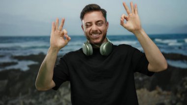 Sakallı ve kulaklıklı genç adam deniz kenarında neşeli bir ifadeyle rahatlamış ve kaygısız bir havayı somutlaştırıyor..