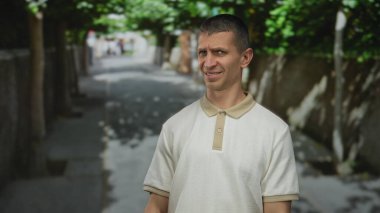 Adam sokakta dikiliyor. Bulanık bir arka planı ve güneşli bir günde yol boyunca uzanan ağaçlar olan günlük kıyafetlerle şaşkın şaşkın bakıyor..