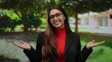 Parkta elleri açık, kırmızı ve siyah bir şekilde gülümseyen bir kadın, genç bir ifade, İspanyol, yeşil gözlüklü, etrafı yeşilliklerle çevrili..