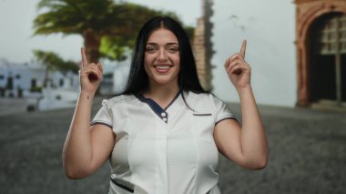 Açık havada gülümseyen bir kadın işaret parmaklarıyla şehrin arka planına doğru bakıyor..