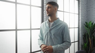 Modern bir ofiste duran genç adam büyük pencerelerin yanında düşünceli görünüyor. Doğal ışık içeri giriyor ve ilham verici bir iş ortamı yaratıyor..