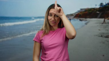Ezik işareti yapan ve güneşli bir sahil sahilinde pembe gömleğiyle kamerayı işaret eden genç bir kadın.