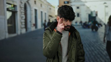 Siyah saçlı ve sakallı genç bir adam. Şehir caddesinde ceket giyiyor. Düşünceli hareketler sergiliyor.