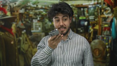 Sakallı, akıllı telefonuyla sesli mesaj kaydeden genç İspanyol bir adam. Kapalı bir pazarda. Etrafı eklektik eşyalarla çevrili ve çizgili gömlek giyiyor..