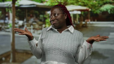 Örgülü saçlı ve gözlüklü bir kadın şehir kafesindeki terasta omuz silkiyor, beyaz gömleğin üzerine gri bir kazak giyiyor, sokak ortamında sıradan bir ifadeyi somutlaştırıyor..