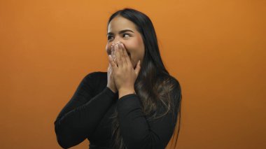 Elleri ağzının üzerinde gülen bir kadın canlı turuncu bir duvarın karşısında neşeli, neşeli bir atmosferi olan, neşeli ve izole bir ortamda neşe ve şaşkınlığı ifade ediyor..