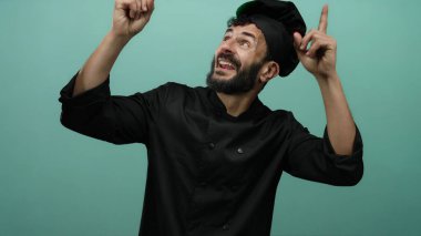 Siyah üniformalı İspanyol erkek aşçı, yeşil bir arka plana doğru bakıyor, coşku ve mutfak uzmanlığını ifade ediyor, neşeyle gülümsüyor..