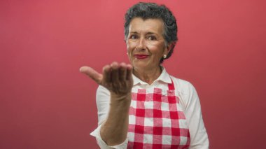 Kırmızı önlüklü, gri saçlı yaşlı bir kadın elini kameraya doğru uzatıyor ve gülümsüyor. Kırmızı bir arka plana doğru. Sıcaklık ve konukseverlik gösteriyor..