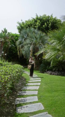 Parlak tropik tatil köyü bahçesinde, güneşli lüks otel ortamında canlı palmiyeler ve egzotik bitkilerle, zarif bir tarzı ve sakin bir atmosferi olan taştan yolda yürüyen bir kadın.