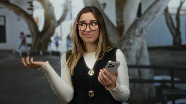 Arka planda ağaçlar, güneşli bir günde siyah yelek ve gözlük takan bir kadın sokakta telefona şaşkın şaşkın bakıyor..