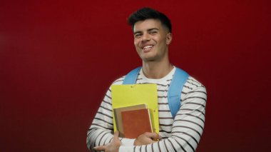 Sırt çantası ve not defterleriyle gülümseyen, parlak kırmızı arka planda, çizgili gömlek giyen, gündelik ve akademik bir stil sergileyen İspanyol genç adam..
