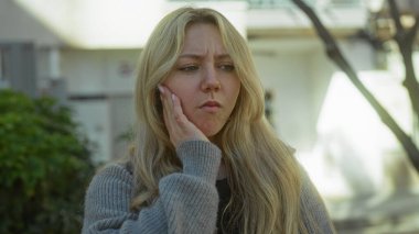 Şehir caddesinde sarışın bir kadın yanağına dokunuyor. Şehir ortamında rahatsız görünüyor. Süveter giyiyor ve duygularını doğal ışıkla ifade ediyor..
