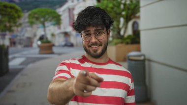 Sakallı genç adam canlı bir sokak ortamında gülümsüyor, gözlük takıyor ve kırmızı çizgili bir gömlek giyiyor, mutluluk ve rahatlama taşıyor..