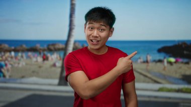 Kırmızı tişörtlü genç Çinli adam kıyı kenarında mavi deniz ve palmiye ağaçlarıyla süslenmiş başparmaklarıyla gülümsüyor ve işaret ediyor. Bu da canlı bir sahil havası olduğunu gösteriyor..