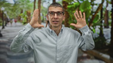 Palmiye ağaçları ile çevrili, şaşırmış ifade ve gözlüklü adam, bir şok ve merak anı yakalar gelişigüzel şehir ortamında.