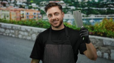 Önlüklü genç adam elinde bir kasap bıçağıyla deniz kenarında geziniyor ve arkasında binalar var..