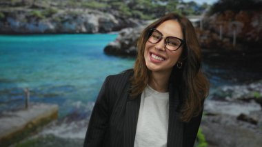Deniz kenarında gülümseyen İspanyol kadın, gözlüklü ve takım elbiseli, açık havada mutluluk ve kendine güveni somutlaştıran, doğal güzellik ve enerji ile deniz kenarına kurulmuş..