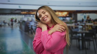 Kadın gülümsüyor ve açık pembe bir tişörtle havaalanında kendine sarılıyor. Arka planı bulanık ve hareketli bir terminal alanı var..
