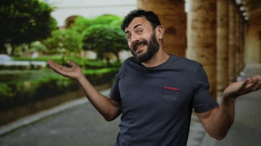 Üniversite kampüsünde sakallı İspanyol bir adam arka planda eski bir mimariyle kafa karışıklığı ya da belirsizliği dile getiriyor..