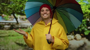 Bahçede gökkuşağı şemsiyeli sarı yağmurluklu genç adam arka planda ağaçlı palmiyeleri gösteriyor.