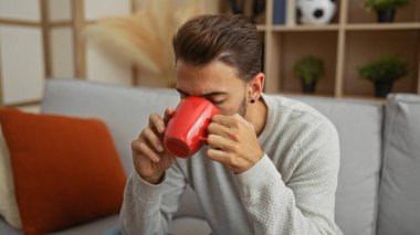 Genç adam, rahat bir oturma odasında, rahat bir kanepe ve sıcak dekor öğeleriyle kahvenin keyfini çıkarıyor. Rahat ve davetkar bir ev atmosferini yakalıyor..