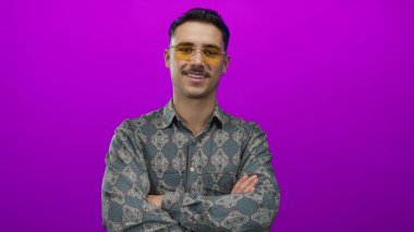 Genç adam kollarını kavuşturarak poz veriyor, parlak pembe arka plana karşı desenli gömlek giyiyor, stüdyo ortamında modern tarz ve hareketli tavırlar sergiliyor.