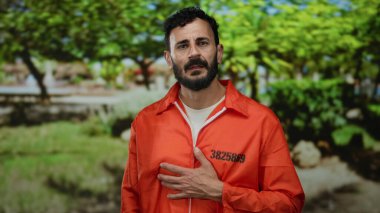 Turuncu hapishane tulumlu İspanyol bir adam canlı yeşil bir parkta ağaçlarla dikiliyor, geniş bir geçmişe sahip, açık havada özgürlük ve neşe öneriyor..