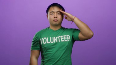 Yeşil gönüllü tişörtü giymiş, kapalı gözlerle selam veren genç adam izole edilmiş mor arka plana karşı duruyor..