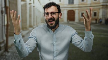 Sakallı İspanyol bir adam, eski bir üniversite kampüsünde şaşkın bir ifade sergiliyor, tarihi mimari ile çevrili, şaşkınlık ve hayal kırıklığı hissi uyandırıyor..