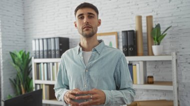 Genç adam, arka planda raflar, klasörler ve bitkilerle modern bir ofis içinde kendine güvenerek duruyor, profesyonel ve yaklaşılabilir bir tavır yakalıyor..