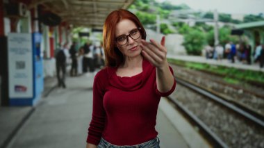 Dışarıdaki tren istasyonu sahnesinde, kırmızı kazaklı bir kadın, tren rayları ve hareketli banliyölerle çevrili, güçlü bir duygu iletiyor..