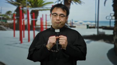 Elleri kelepçeli papaz dışarıda duruyor. Arka planda kırmızı çubuklar ve palmiye ağaçları var.